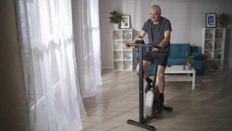 Der-Grauhaarige-Mann-Trainiert-Im-Wohnzimmer-Auf-Dem-Spinning-Bike-Einen-Gesunden-Lebensstil-Und-Hält-Im-Mittleren-Alter-Eine-Gute-Körperliche-Verfassung